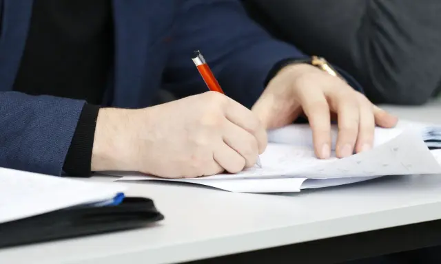 Ręka pisze długopisem po papierze, przygotowując przykładowy artykuł po angielsku na maturę.