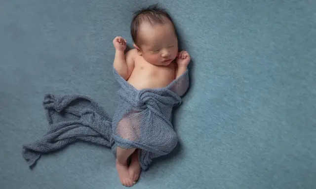 Neonato dorme avvolto in un telo blu, con le braccia sollevate e i pugni chiusi.