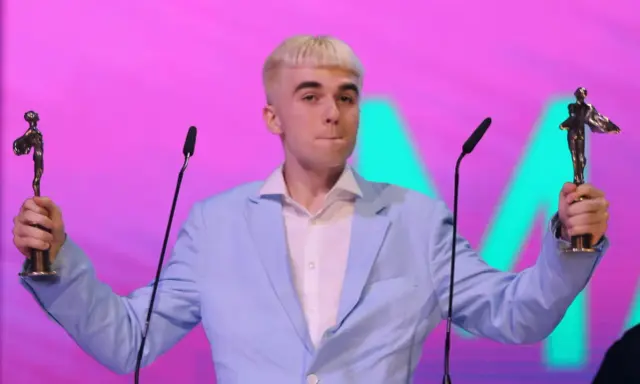 Mc Mata z dwoma statuetkami na scenie, w jasnoniebieskiej marynarce i białej koszuli.
