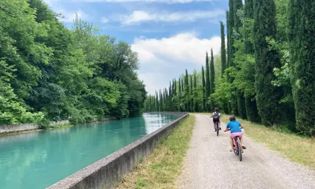 Bambini in bicicletta lungo la ciclovia del Mincio, con il canale turchese e filari di cipressi.