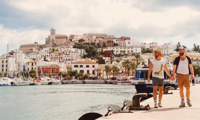 A gay couple walks hand-in-hand along a harbor in Spain, with a white-washed town and ancient fortress rising behind them.