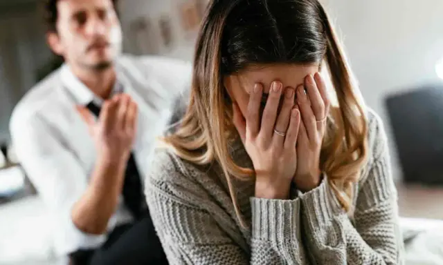 Kobieta zakrywa twarz dłońmi, płacząc. Mężczyzna w białej koszuli gestykuluje, sugerując przemoc w białych rękawiczkach.