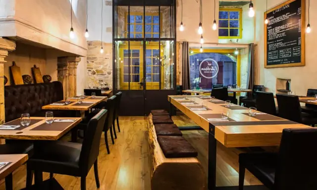 Ambiance chaleureuse au restaurant "Les Fougeres". Tables en bois brut, banquette en cuir et cheminée imposante créent un décor rustique et élégant.