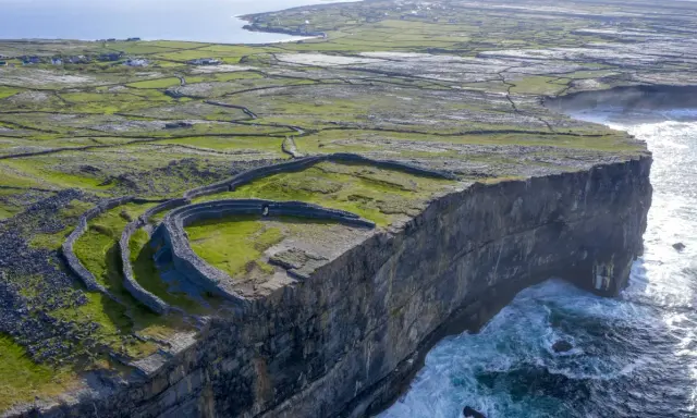 Steile Klippen und alte Steinmauern auf einer grünen Insel vor Galway, Irland. Wellen schlagen gegen die Felsen.