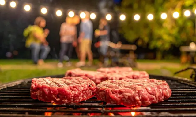 Frische Burger-Patties liegen auf dem Grill. Im Hintergrund feiern Freunde unter Lichterketten. So macht man Hackfleisch selber für den perfekten Grillabend.
