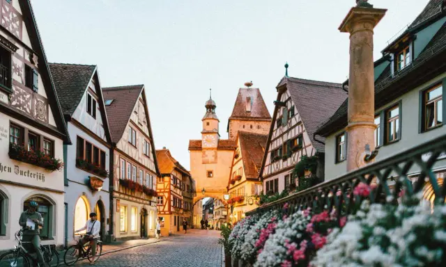 A charming street in gay Germany, with historic buildings, cobblestones, and flowers. Two people on bikes pause to take in the view.