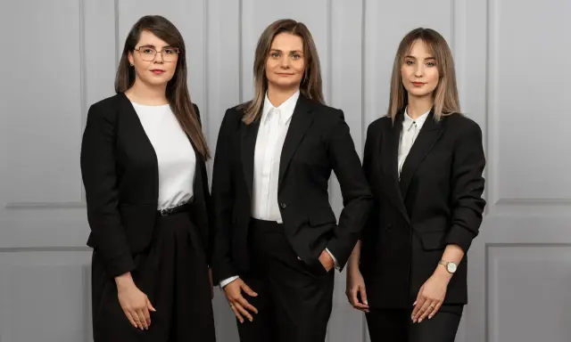 Trzy kobiety w czarnych garniturach stoją przed szarą ścianą.