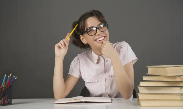 Ragazza con occhiali e matita gialla, un libro aperto e pile di libri, un'espressione pensierosa che ricorda l'anastrofe figura retorica.