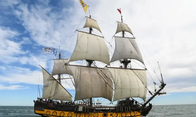 Le Shtandart, bateau historique aux voiles blanches, navigue sur une mer agitée sous un ciel nuageux.
