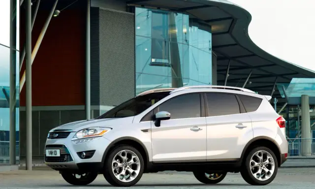 Biały Ford Kuga 2010 zaparkowany przed nowoczesnym budynkiem z przeszkloną fasadą.