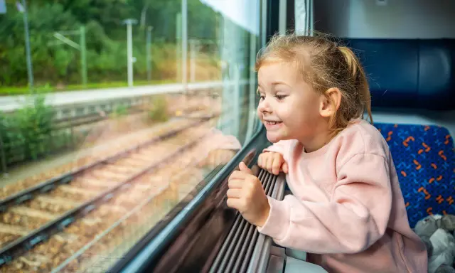 Uśmiechnięta dziewczynka patrzy przez okno pociągu. Zastanawia się, od ilu lat można jeździć samemu pociągiem, podziwiając mijane krajobrazy.