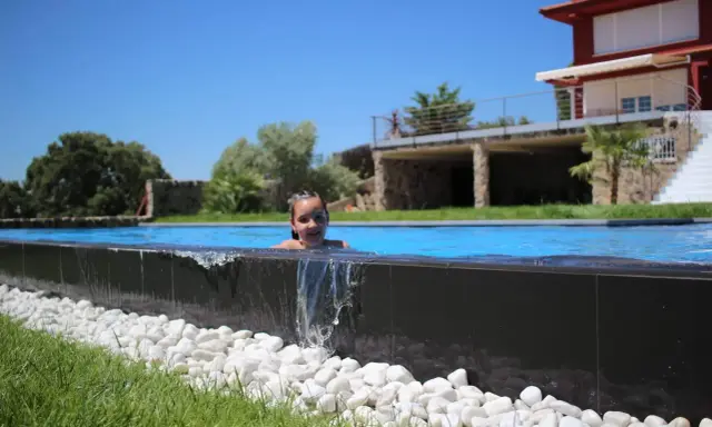 Niña sonriente en una piscina desbordante, con agua cayendo sobre piedras blancas y césped verde. Al fondo, una casa moderna y árboles frondosos bajo un cielo azul.