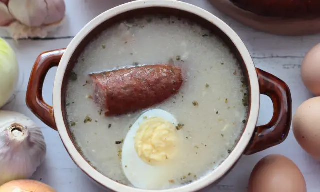 Aromaticzny zakwas na żurek w glinianym naczyniu, z kiełbasą i jajkiem. Obok czosnek, cebula i jajka.