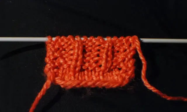 En orange stickad provlapp med ett mönster av vriden resår på en sticka. En lös tråd hänger ner.