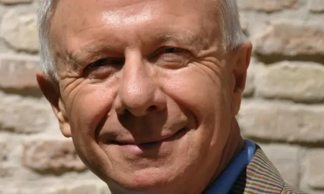 Primo piano sorridente di padre Alberto Maggi, con rughe d'espressione e capelli grigi, sullo sfondo di un muro di mattoni.