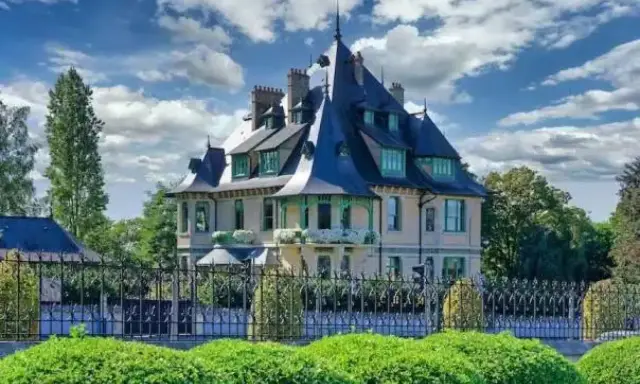 La villa Demoiselle, une demeure de caractère avec ses toits pointus et sa façade claire, se dresse fièrement derrière une grille ouvragée et des buissons taillés.