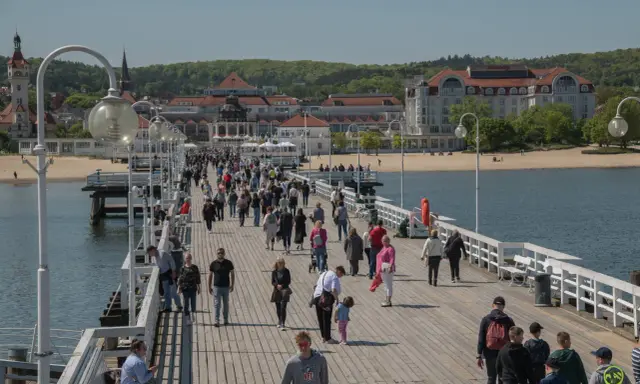 Tłum ludzi spaceruje po molo w Sopocie, jednym z najpopularniejszych gdańsk atrakcje. W tle widać plażę i zabytkowe budynki.