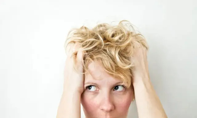 Kobieta z blond włosami wplata dłonie we włosy, jej wyraz twarzy sugeruje wahania nastroju i niepokój.
