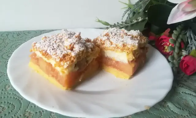 Pyszna szarlotka z bezą i kruszonką, z jabłkami i cukrem pudrem, na białym talerzu, obok kwiatów.