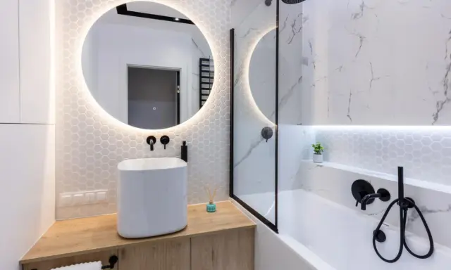 Elegante bagno senza finestra con lavabo bianco, specchio rotondo illuminato e doccia con finiture nere.
