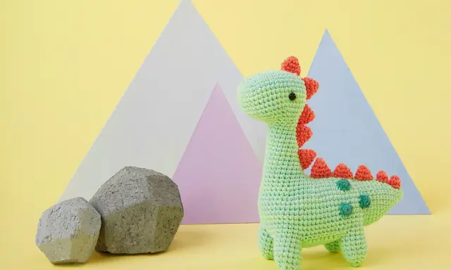 Adorable dinosaurio crochet verde con púas naranjas y detalles verdes. Dos rocas grises y formas geométricas de colores pastel al fondo.