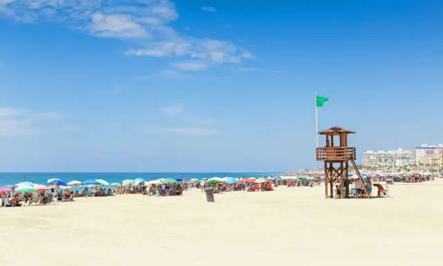 Sonniger Strand in Rota, Spanien, mit vielen bunten Sonnenschirmen und Menschen. Ein Rettungsschwimmerturm mit grüner Flagge steht am Meer.