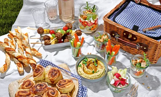 Leckere Picknick-Snacks: Fleischbällchen-Spieße, Gebäckstangen, Hummus, Gemüsesticks und süße Schnecken auf einer Decke.