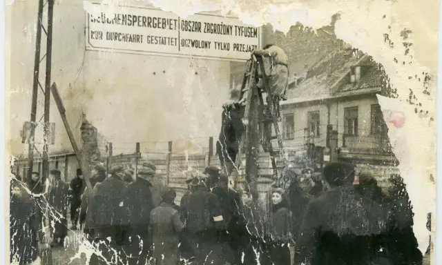 1941. Ludzie stoją przed znakiem "Obszar zagrożony tyfusem". W tle budynki i druty kolczaste.