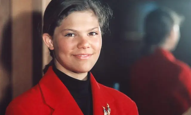 Kronprinsessan Victoria ung, med ett varmt leende, bär en röd kavaj och en svart polotröja. En vacker brosch pryder kavajen.