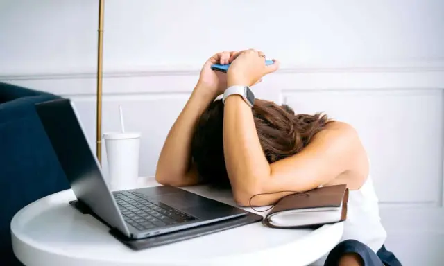 Personne épuisée, tête sur les bras, téléphone à la main, devant un ordinateur portable. L'effet écran de la fatigue est palpable.