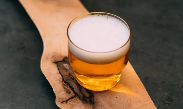 Szklanka złocistego piwa z białą pianką, prezentująca standardową dawkę alkoholu, stoi na drewnianej desce.