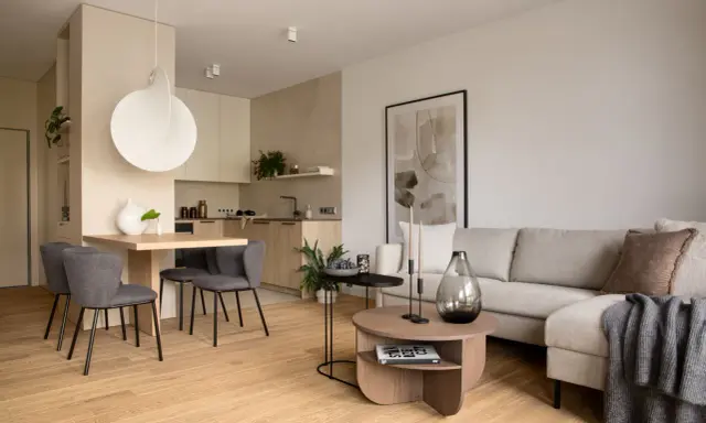 Beżowy salon inspiracje: przytulna przestrzeń z szarą sofą, drewnianym stołem i nowoczesną kuchnią.