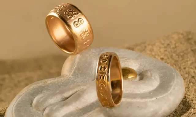 Due anelli d'oro con incisioni uniche, che mostrano diversi tipi di fedi nuziali, su una pietra a forma di mano con sabbia.