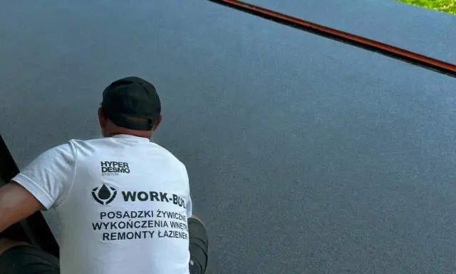 Pracownik w koszulce z napisem "POSADZKI ŻYWICZNE" wykonuje prace na ciemnej powierzchni.