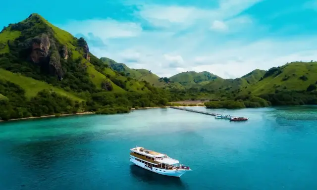 Un'isola lussureggiante con colline verdi e un mare turchese, dove si trova Bali, con una nave da crociera che naviga nelle acque cristalline.