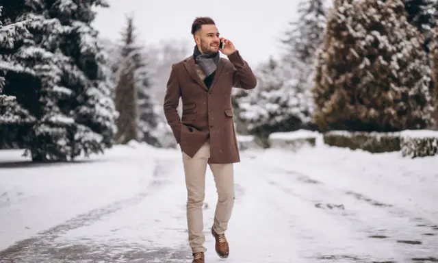 Man in stylish winter cocktail attire for men, wearing a brown coat and scarf, walks through a snowy park while on the phone.