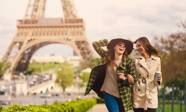 Due amiche ridono passeggiando a Parigi, con la Torre Eiffel sullo sfondo. Perfetto per capire cosa mettere in valigia per Parigi: cappotti leggeri, cappello e occhiali da sole.
