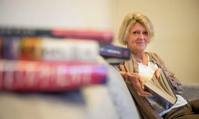 En kvinna med blont hår sitter med en bok i handen, omgiven av fler böcker. Hon ler mot kameran, som om hon delar en hemlighet från de fenomenala fruntimren på Grand Hotel.