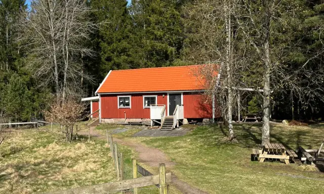 Rött torp med orange tak, omgivet av skog. En stig leder fram till torpet Freden, där picknickbord står redo för besökare.