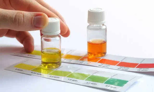 Mano sosteniendo un vial con líquido amarillo sobre tiras de prueba de color para saber la dureza del agua de mi zona. Otro vial con líquido naranja al lado.