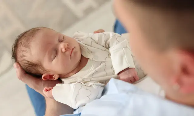Un neonato dorme sereno tra le braccia di un genitore, cullato da suoni che ricordano i rumori bianchi, creando un'atmosfera di pace.