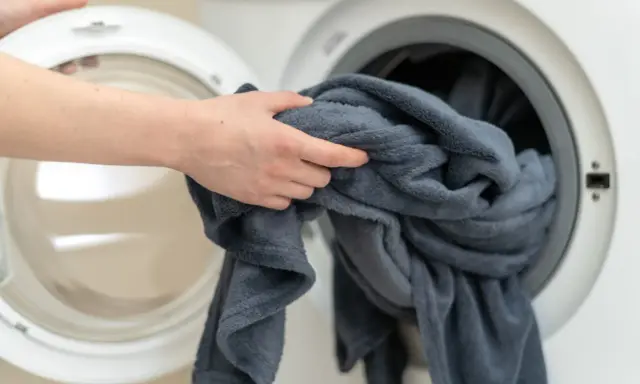 Ręka wkłada szary koc do pralki. Tak jak prac koce, by były czyste i pachnące.