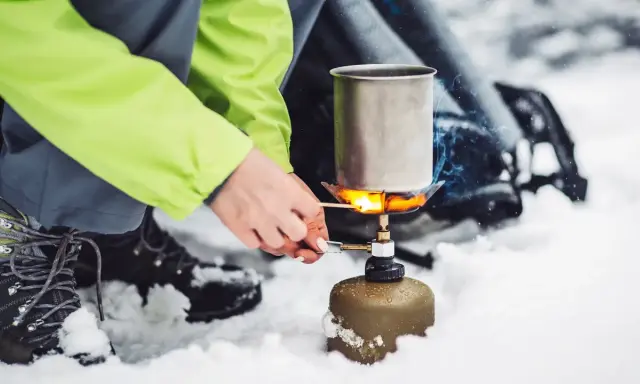 Tänd ett bensinkök i snön för att värma upp en kastrull. En person i en ljusgrön jacka och vandringskängor förbereder mat utomhus.