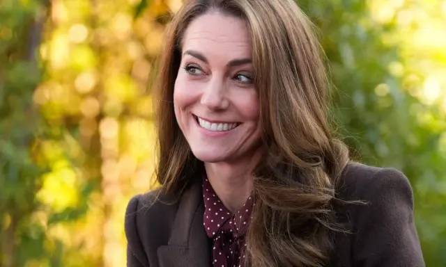 Kate Middleton sonríe con calidez, con su cabello castaño cayendo en cascada. El fondo bokeh de tonos otoñales complementa su elegante atuendo.