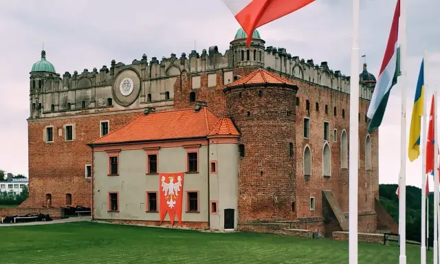 Majestatyczny zamek golubski z polską flagą i innymi flagami narodowymi na trawiastym dziedzińcu.