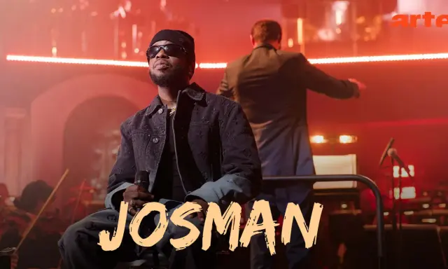 Josman, francuski rap artysta, siedzi na scenie z mikrofonem, w tle orkiestra i dyrygent.