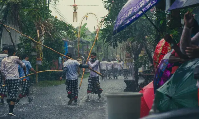 Ulewny deszcz na Bali podczas pory deszczowej. Mężczyźni w tradycyjnych strojach niosą drewniane kije, a widzowie chronią się pod kolorowymi parasolami.