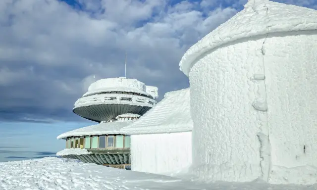 Zimowe krajobrazy na Śnieżce. Budynki obserwatorium pokryte lodem i śniegiem, niebo z chmurami.