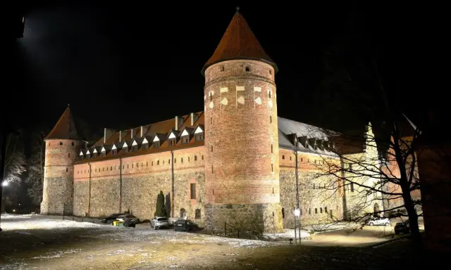 Zamek w Bytowie nocą, oświetlony, z wieżami i kamiennymi murami.