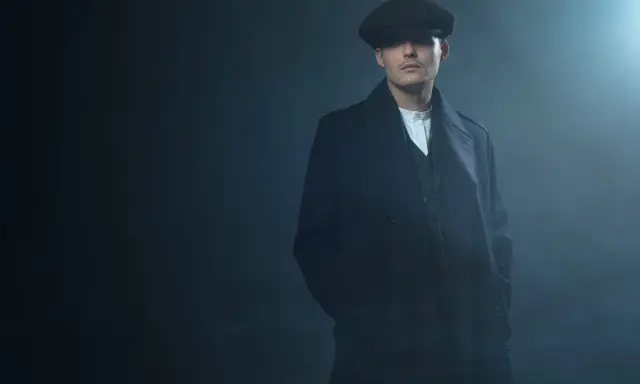 Młody mężczyzna w czapce i płaszczu, z charakterystyczną fryzurą w stylu Peaky Blinders, stoi w zadymionym mroku.